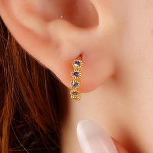 Puede incluir: Aretes de aro dorados con pequeñas piedras preciosas púrpuras dispuestas en un patrón circular.
