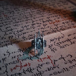 Pu&ograve; includere: Un anello color argento con un design intricato, caratterizzato da un emblema centrale e punte verticali. L'anello &egrave; posto su una pergamena invecchiata coperta di testo manoscritto e segni rossi, creando un'estetica gotica o fantasy.