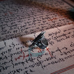 Pu&ograve; includere: Un anello d'argento dal design dettagliato, appoggiato su una pagina ricoperta di testo manoscritto. L'anello ha un aspetto unico e strutturato. La pagina presenta dettagli rossi e neri.