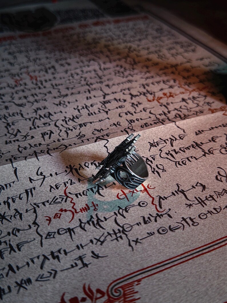 Pu&ograve; includere: Un anello d'argento dal design dettagliato, appoggiato su una pagina ricoperta di testo manoscritto. L'anello ha un aspetto unico e strutturato. La pagina presenta dettagli rossi e neri.