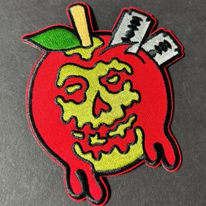 May include: A red apple with a dripping green and yellow skull inside. The apple has a green leaf and a brown stick on top. There are two silver razor blades sticking out of the top of the apple.