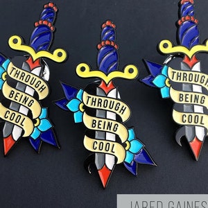 Puede incluir: Tres pines de esmalte que representan una daga estilizada con un mango amarillo y una hoja azul. Cada pin tiene una pancarta amarilla envuelta alrededor de la daga con el texto "Through Being Cool" en negro.