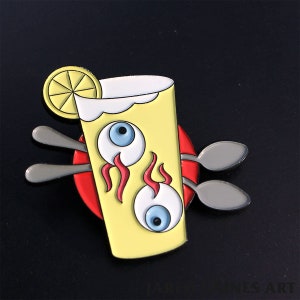 May include: A yellow enamel pin of a glass of lemonade with two eyeballs and flames. The glass has a lemon wedge on the rim and two spoons are behind it.