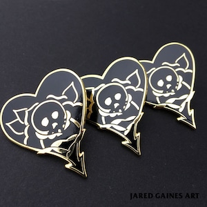 Puede incluir: Tres pines negros en forma de corazón con contornos dorados. Cada pin presenta una calavera blanca dentro de una rosa. El texto "JARED GAINES ART" es visible en la parte inferior de la imagen.