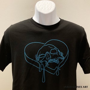 Puede incluir: Camiseta negra con un contorno azul de un coraz&oacute;n con una calavera en su interior. El coraz&oacute;n gotea un l&iacute;quido azul. El texto "JARED GAINES ART" est&aacute; en la parte inferior de la camiseta.