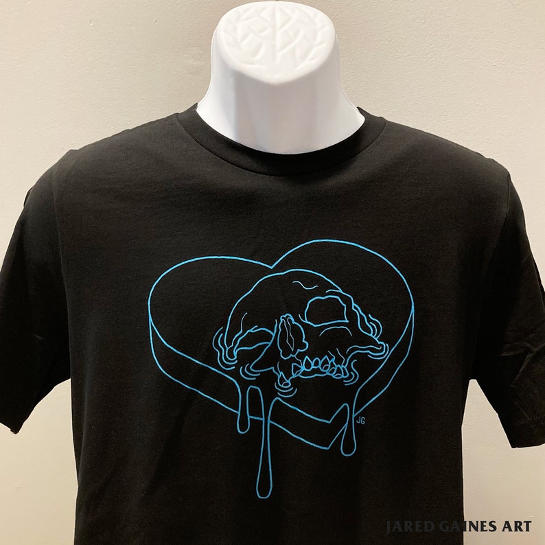 Puede incluir: Camiseta negra con un contorno azul de un coraz&oacute;n con una calavera en su interior. El coraz&oacute;n gotea un l&iacute;quido azul. El texto "JARED GAINES ART" est&aacute; en la parte inferior de la camiseta.