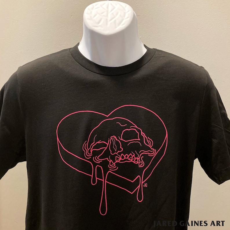 Puede incluir: Camiseta negra con un contorno rosa de una calavera dentro de un coraz&oacute;n que gotea. El texto "JG" est&aacute; en la esquina inferior derecha del coraz&oacute;n. El texto "JARED GAINES ART" est&aacute; en la parte inferior de la camiseta.