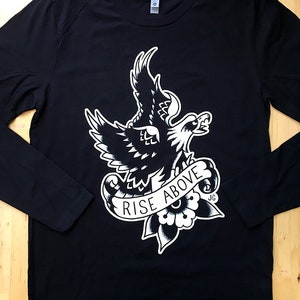 Puede incluir: Camiseta negra de manga larga con un gráfico blanco de un águila con una pancarta que dice "Rise Above".