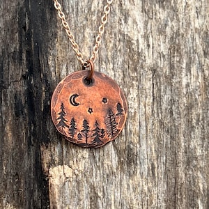 May include: A copper necklace with a round pendant featuring a crescent moon, stars, and a silhouette of trees. The pendant is engraved with a woodland scene.