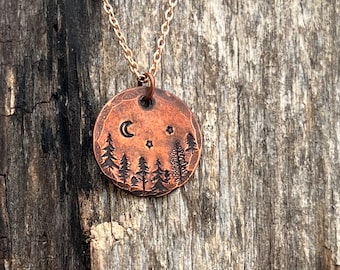 May include: A copper necklace with a round pendant featuring a crescent moon, stars, and a silhouette of trees. The pendant is engraved with a woodland scene.