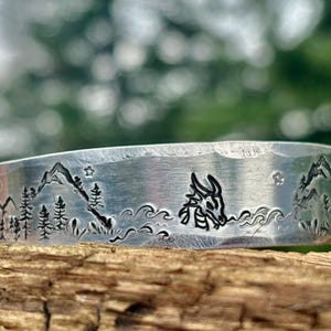 May include: A silver-colored cuff bracelet with a mountain and forest design. The bracelet features a dragon's head and a landscape with trees, mountains, and stars. The bracelet is resting on a piece of wood.