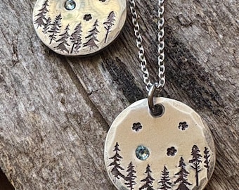 May include: Two silver pendant necklaces. Each pendant is a hammered, circular disc with a forest scene etched into it. The design includes trees, stars, and a small, round, blue gemstone. The necklaces hang on silver chains.