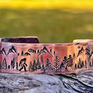 May include: A hammered copper cuff bracelet featuring a dark etched landscape design. The engraving depicts a silhouette of a creature resembling Bigfoot walking through a dense forest of pine trees with mountain peaks and small stars in the background.