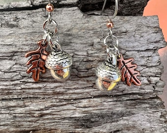 May include: A pair of dangle earrings featuring silver-toned acorn charms with textured caps and copper-colored oak leaf charms. The earrings have silver-colored ear wires and small copper-colored beads.