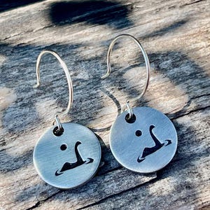 May include: A pair of silver earrings with a circular design featuring a silhouette of a Loch Ness Monster. The earrings are hanging from silver hooks.