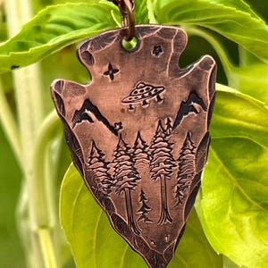 May include: A copper arrowhead pendant necklace with a detailed design of a forest, mountains, and a UFO. The pendant has a rustic, textured finish.