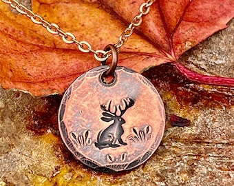 May include: A copper necklace with a round pendant featuring a stylized image of a deer with antlers. The pendant is textured and has a rustic look.
