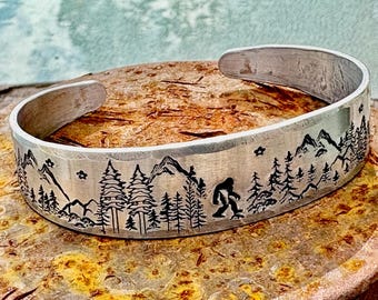 May include: Silver-toned cuff bracelet with a mountain and forest design. The bracelet features a Bigfoot silhouette, trees, mountains, and stars. The cuff is approximately 2.5 inches wide.