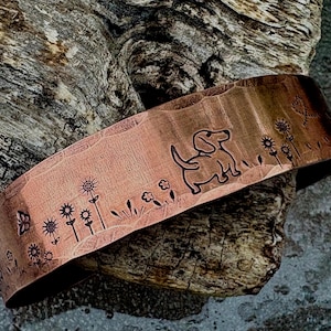 May include: A wide copper cuff bracelet featuring a hammered texture and stamped designs. The metal surface displays a line art illustration of a small dog, several stylized flowers, and a butterfly, all etched into the warm-toned copper material.