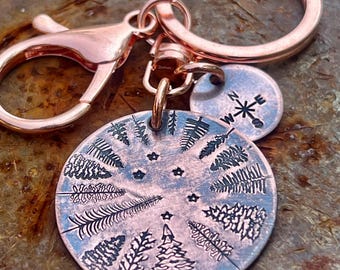 May include: A copper-colored keychain with a round pendant featuring etched trees and stars. A smaller, circular tag with a compass rose is attached. The keychain includes a lobster clasp and a split ring.