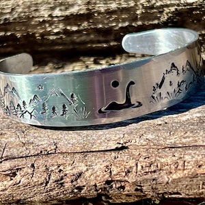 May include: A silver bracelet with an engraved design of a mountain range and a mythical creature, possibly a Loch Ness Monster, in a lake. The bracelet is on a wooden surface.