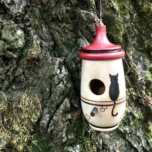 Puede incluir: Un comedero para p&aacute;jaros de madera con techo rojo y silueta de gato negro. El comedero est&aacute; colgado de un &aacute;rbol y tiene un agujero de entrada redondo. El comedero est&aacute; pintado con detalles en negro y marr&oacute;n.