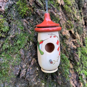 May include: A decorative wooden birdhouse with a red roof and a painted hummingbird design. The birdhouse is cylindrical with a round entrance hole and is hanging from a metal hook. The background is a tree trunk covered in moss.