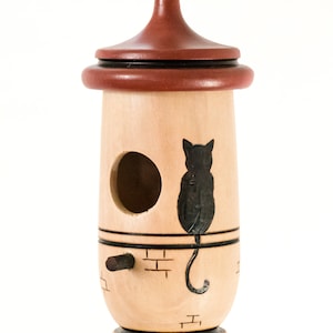 Puede incluir: Una casa para p&aacute;jaros de madera con cuerpo marr&oacute;n claro y techo marr&oacute;n oscuro. Una silueta de gato negro est&aacute; pintada en el lateral. La casa para p&aacute;jaros tiene un agujero de entrada redondo y una peque&ntilde;a percha.