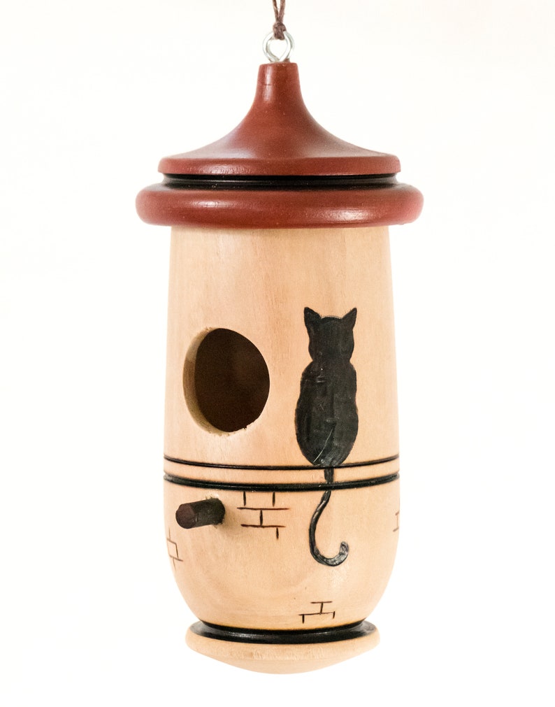 Puede incluir: Una casa para p&aacute;jaros de madera con cuerpo marr&oacute;n claro y techo marr&oacute;n oscuro. Una silueta de gato negro est&aacute; pintada en el lateral. La casa para p&aacute;jaros tiene un agujero de entrada redondo y una peque&ntilde;a percha.