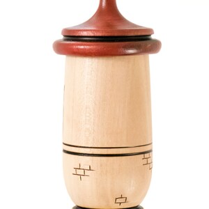 Puede incluir: Un adorno de madera con un cuerpo marr&oacute;n claro y una parte superior roja oscura. El adorno tiene forma cil&iacute;ndrica con un patr&oacute;n decorativo de ladrillos grabado en la madera. Una fina cuerda de cuero est&aacute; unida a la parte superior para colgar.