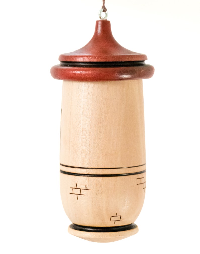 Puede incluir: Un adorno de madera con un cuerpo marr&oacute;n claro y una parte superior roja oscura. El adorno tiene forma cil&iacute;ndrica con un patr&oacute;n decorativo de ladrillos grabado en la madera. Una fina cuerda de cuero est&aacute; unida a la parte superior para colgar.