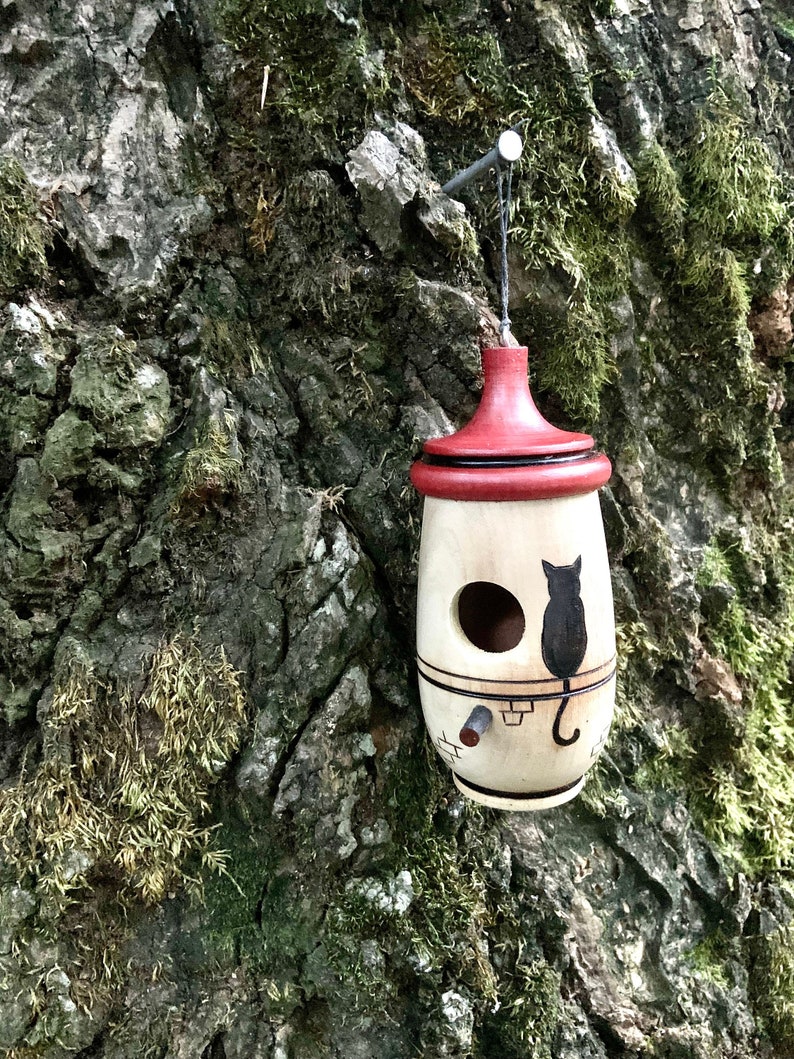 Puede incluir: Un comedero para p&aacute;jaros de madera con techo rojo y silueta de gato negro. El comedero est&aacute; colgado de un &aacute;rbol y tiene un agujero de entrada redondo. El comedero est&aacute; pintado con detalles en negro y marr&oacute;n.