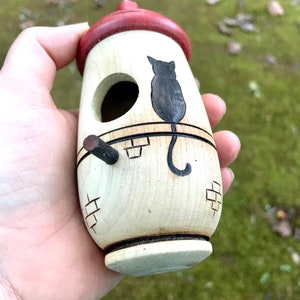 Puede incluir: Una casa para p&aacute;jaros de madera con techo rojo y silueta de gato negro. La casa para p&aacute;jaros es cil&iacute;ndrica con una peque&ntilde;a percha y un agujero de entrada circular. El dise&ntilde;o incluye tallas en forma de ladrillo y se sostiene en una mano.