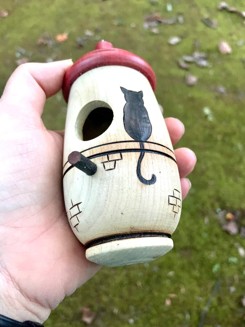 Puede incluir: Una casa para p&aacute;jaros de madera con techo rojo y silueta de gato negro. La casa para p&aacute;jaros es cil&iacute;ndrica con una peque&ntilde;a percha y un agujero de entrada circular. El dise&ntilde;o incluye tallas en forma de ladrillo y se sostiene en una mano.