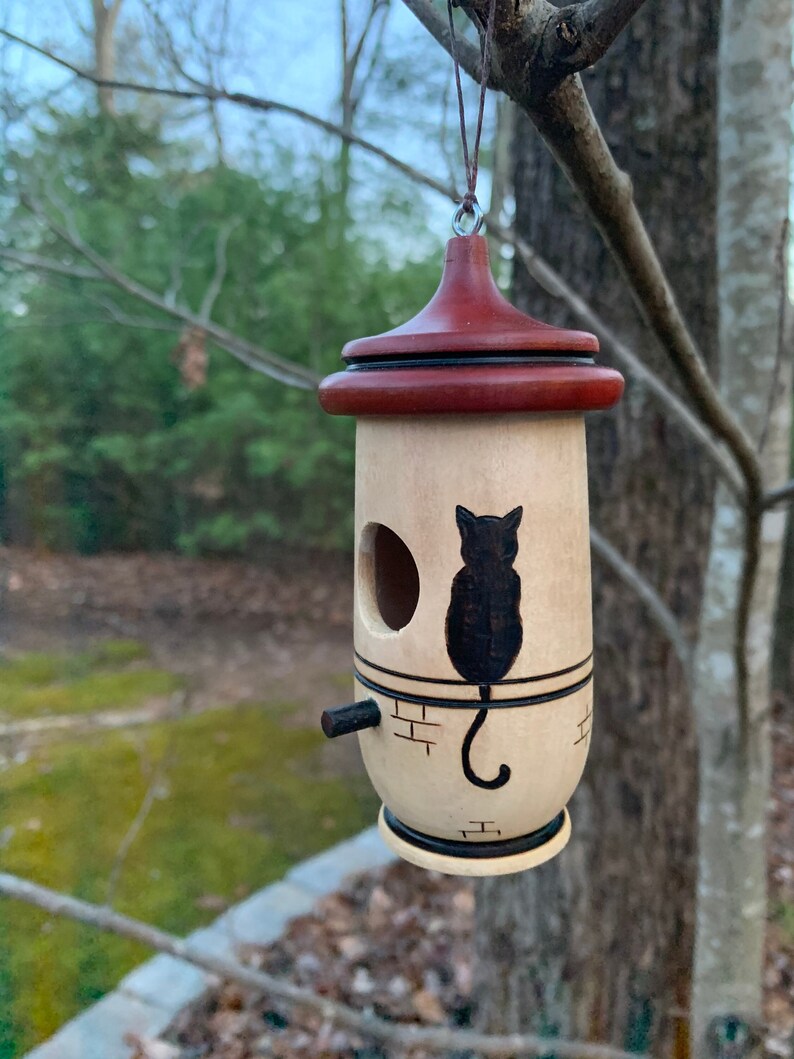 Puede incluir: Una casita para p&aacute;jaros de madera con cuerpo cil&iacute;ndrico y techo c&oacute;nico rojo oscuro. Una silueta de gato negro est&aacute; pintada en la parte delantera. La casita tiene una peque&ntilde;a percha y una entrada circular. Cuelga de un hilo fino.