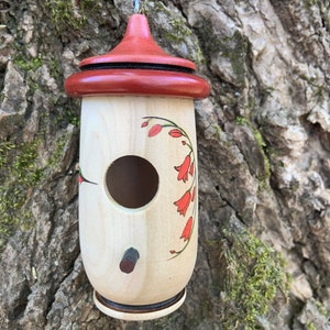 May include: A wooden birdhouse with a red roof and decorative floral design. The birdhouse is cylindrical with a round entry hole and a small perch. It is hanging from a thin wire against a tree trunk.