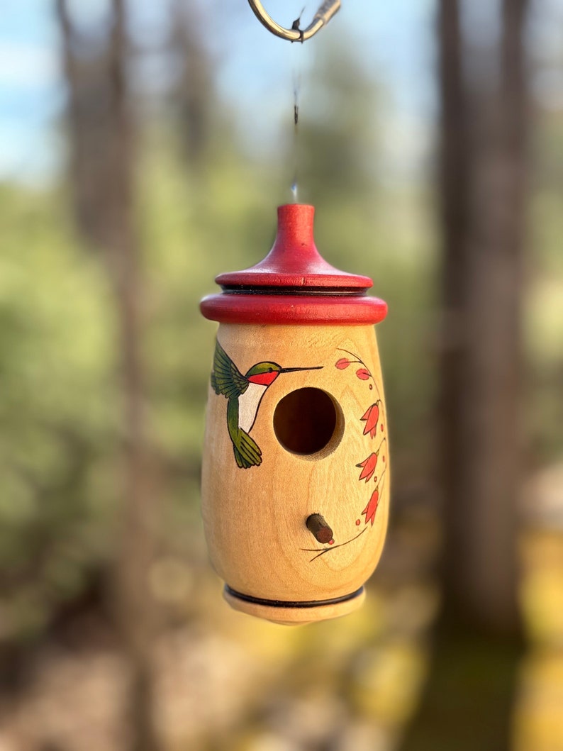Image of a very small wooden hummingbird House. The bottom of the house is round with a colored image of a hummingbird on the left side of the hole and perch. On the right side is a red flowering vine. The top is a red colored wood, also round.