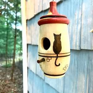 Puede incluir: Un comedero para p&aacute;jaros colgante con cuerpo crema y parte superior roja. Presenta una silueta de gato negro y una entrada circular. Se observa una peque&ntilde;a percha. El comedero est&aacute; suspendido de una cuerda negra.