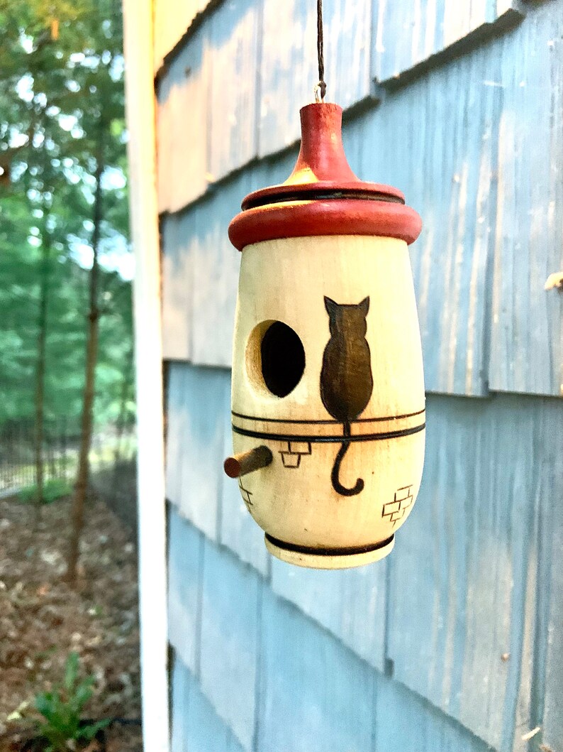 Puede incluir: Un comedero para p&aacute;jaros colgante con cuerpo crema y parte superior roja. Presenta una silueta de gato negro y una entrada circular. Se observa una peque&ntilde;a percha. El comedero est&aacute; suspendido de una cuerda negra.