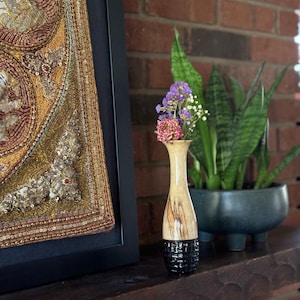 May include: A small, decorative vase with a light-colored top and a dark, textured base. The vase holds a small bouquet of dried flowers in shades of purple, pink, and white. The vase is displayed on a wooden shelf with a framed artwork and a potted plant.