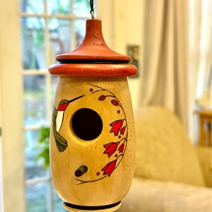 May include: A wooden birdhouse with a red-brown top and a light-colored body. The birdhouse features a painted hummingbird and red flower design. A small hole is visible for entry. The birdhouse is hanging from a black string.