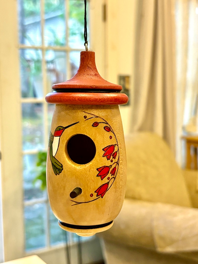 May include: A wooden birdhouse with a red-brown top and a light-colored body. The birdhouse features a painted hummingbird and red flower design. A small hole is visible for entry. The birdhouse is hanging from a black string.