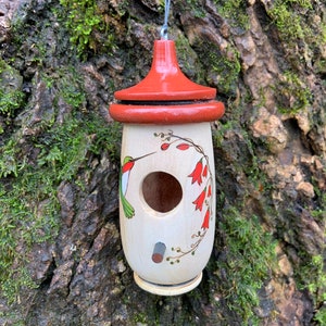 May include: A wooden birdhouse with a red roof and a cream-colored body. The birdhouse is decorated with a hummingbird and red flower illustrations. It hangs from a metal wire against a mossy tree trunk.