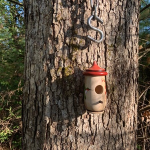May include: A wooden birdhouse with a red roof and a round entry hole hangs from a metal chain attached to a tree. The birdhouse is cylindrical with painted floral and hummingbird designs. The tree bark is textured and brown.