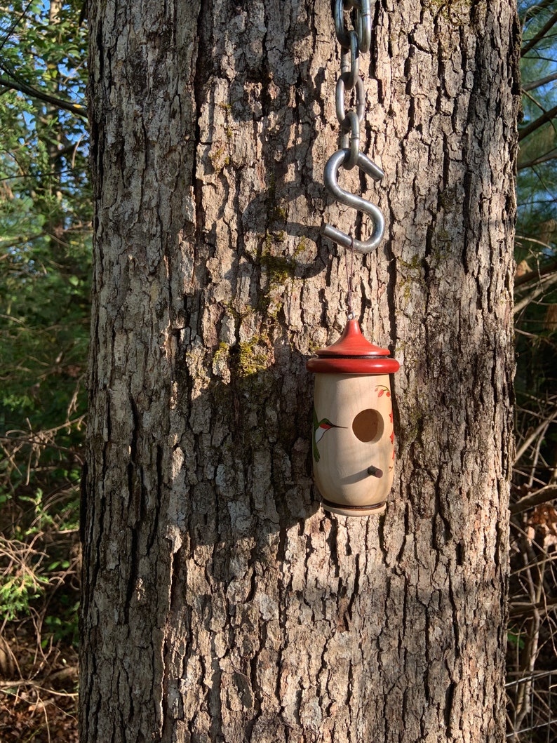 May include: A wooden birdhouse with a red roof and a round entry hole hangs from a metal chain attached to a tree. The birdhouse is cylindrical with painted floral and hummingbird designs. The tree bark is textured and brown.