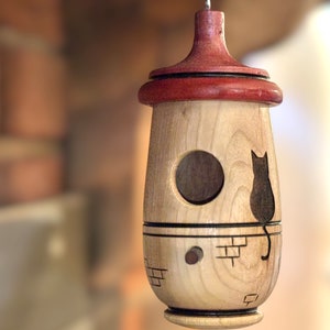 Puede incluir: Una pajarera de madera con un agujero de entrada redondo y una silueta de gato. La pajarera tiene una parte superior roja y un acabado de madera clara. Cuelga de un gancho de metal.