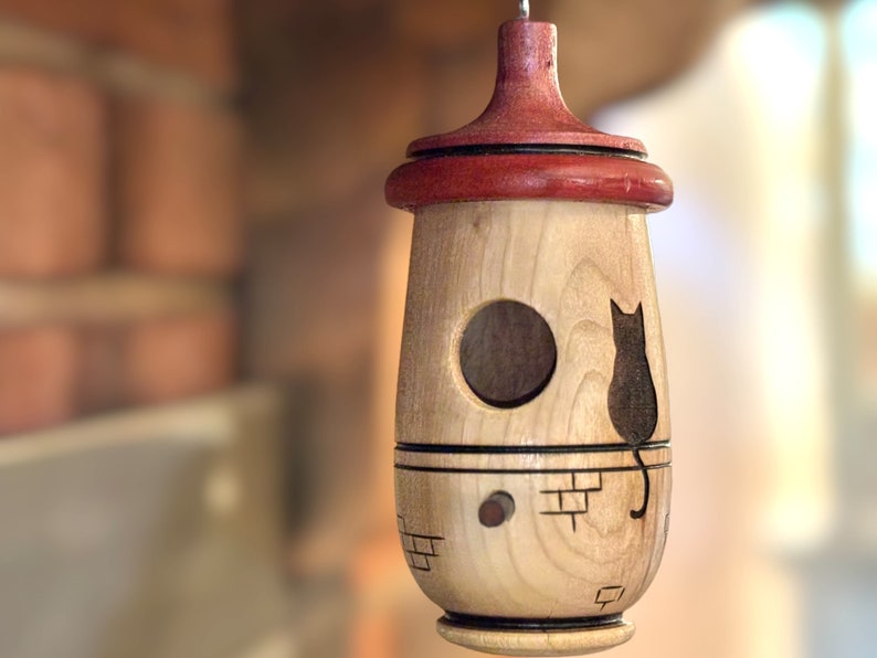 Puede incluir: Una pajarera de madera con un agujero de entrada redondo y una silueta de gato. La pajarera tiene una parte superior roja y un acabado de madera clara. Cuelga de un gancho de metal.