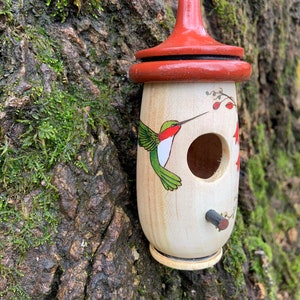 May include: A wooden birdhouse with a red top and a hummingbird illustration. The birdhouse is cylindrical with a small perch and a round entry hole. The hummingbird is depicted with green wings and a red throat. The birdhouse hangs from a thin string.