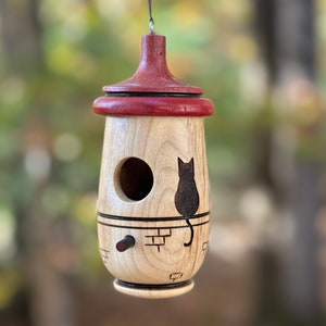 Puede incluir: Una casa para p&aacute;jaros de madera con un cuerpo de madera clara y una parte superior roja oscura. Una silueta de gato negro est&aacute; grabada en el lateral. La casa para p&aacute;jaros tiene un agujero de entrada redondo y una peque&ntilde;a percha. Cuelga de un hilo negro fino.