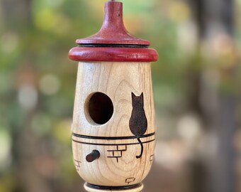 May include: A wooden birdhouse with a light wood body and a dark red top. A black cat silhouette is etched on the side. The birdhouse has a round entry hole and a small perch. It hangs from a thin black string.